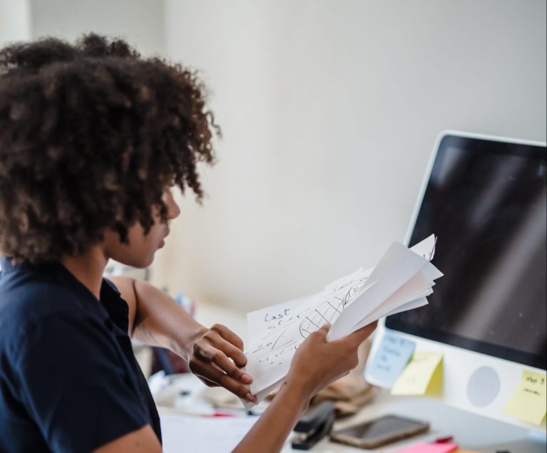 Imagem de uma mulher sentada em frente a um computador no qual há post-its colados na tela. Ela segura papéis referentes à análise de um projeto do qual ela participa.