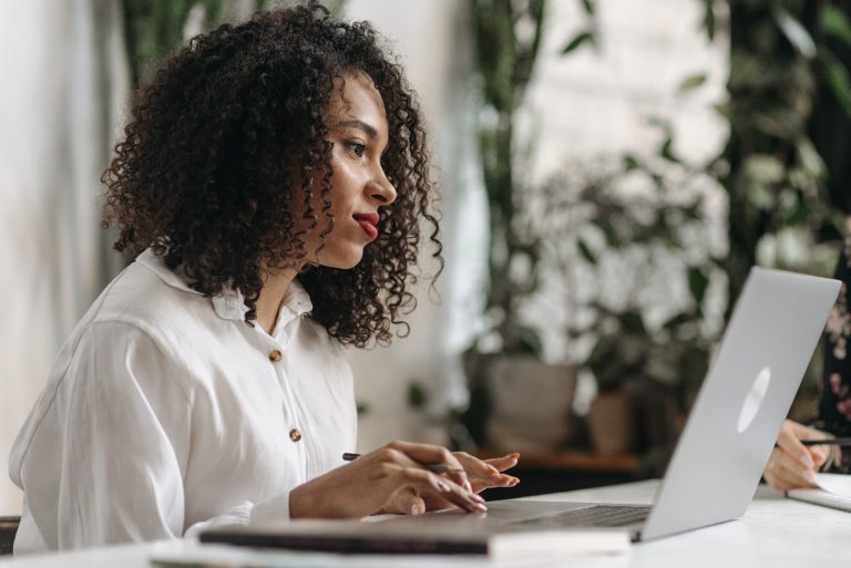 Imagem de uma mulher. Ela tem cabelos cacheados, usa uma camisa branca e manuseia um notebook. A foto foi escolhida para ilustrar a nota sobre o Matchfunding Novos Caminhos.